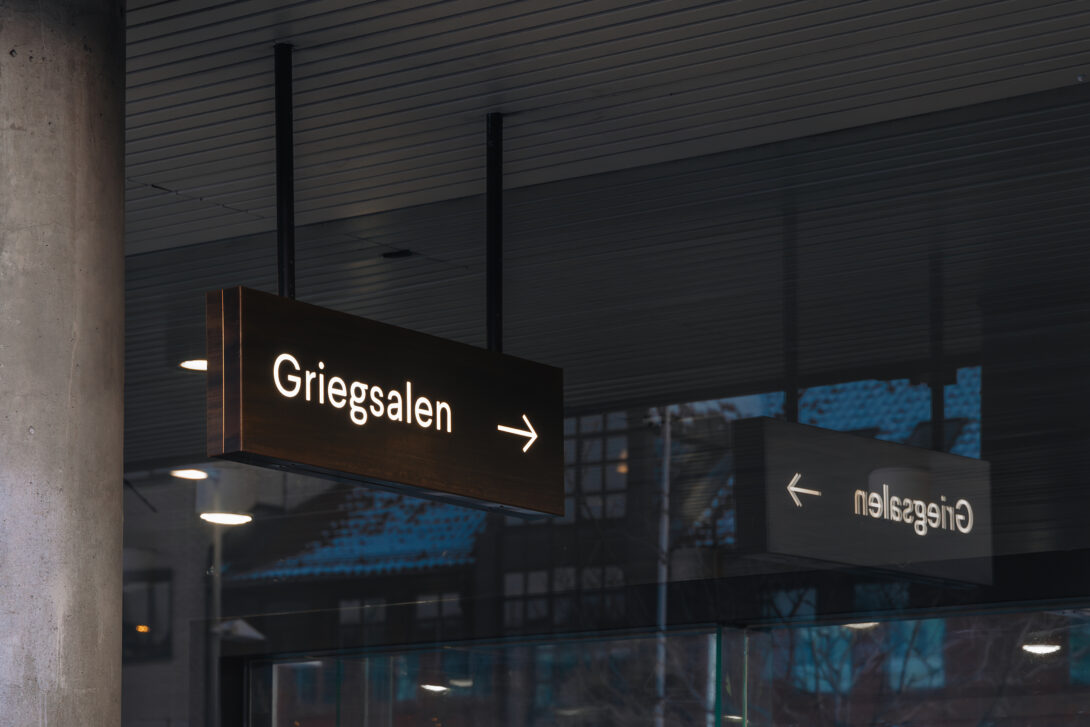 Belyst skilt med teksten Griegsalen i taket med refleksjon i glassveggen