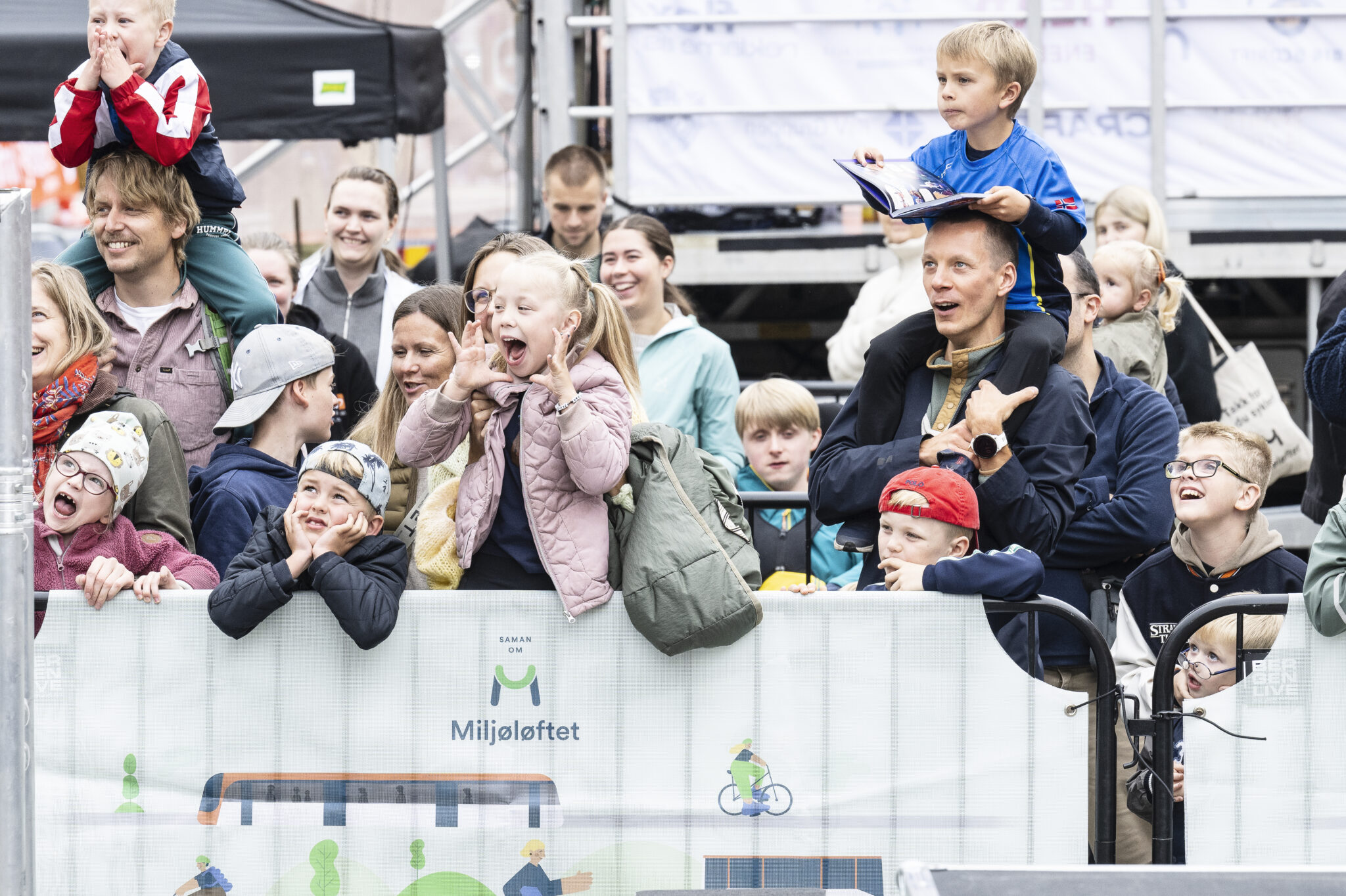 Barn og voksne samlet tett langs en arena under Mobilitetsveka. Publikum reagerer engasjert på aktiviteten foran dem, mens en illustrert Miljøløftet-profil danner en lys visuell flate i forgrunnen.