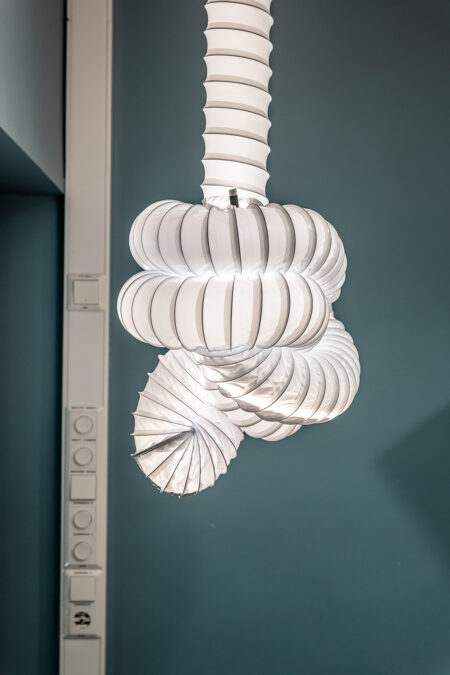 Skulpturell lampe av ventilasjonsrør som henger foran en petroleumsfarget vegg i biblioteket.