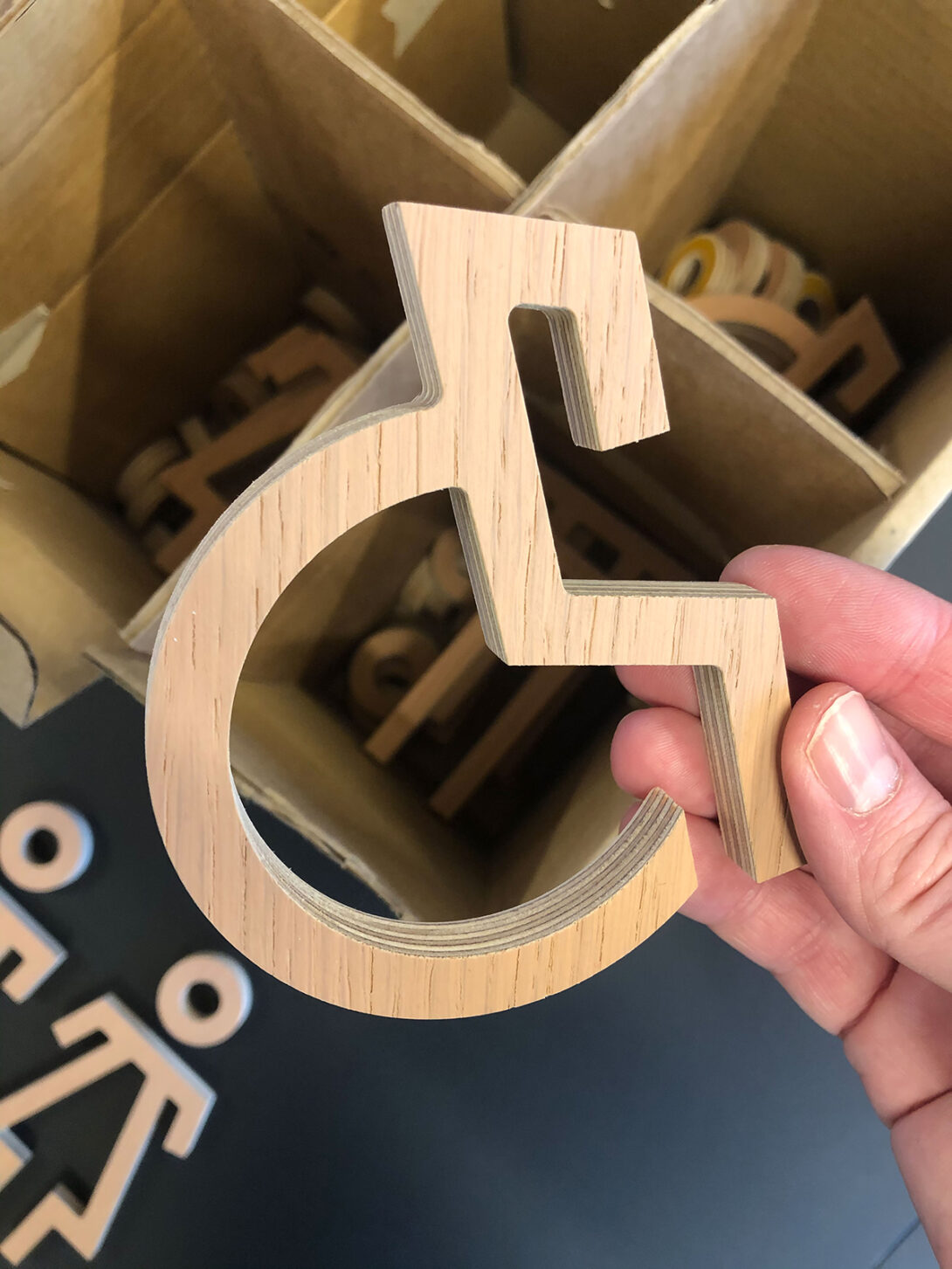 En hånd som holder opp et utskåret rullestolsymbol i tre for skilt og merking.
